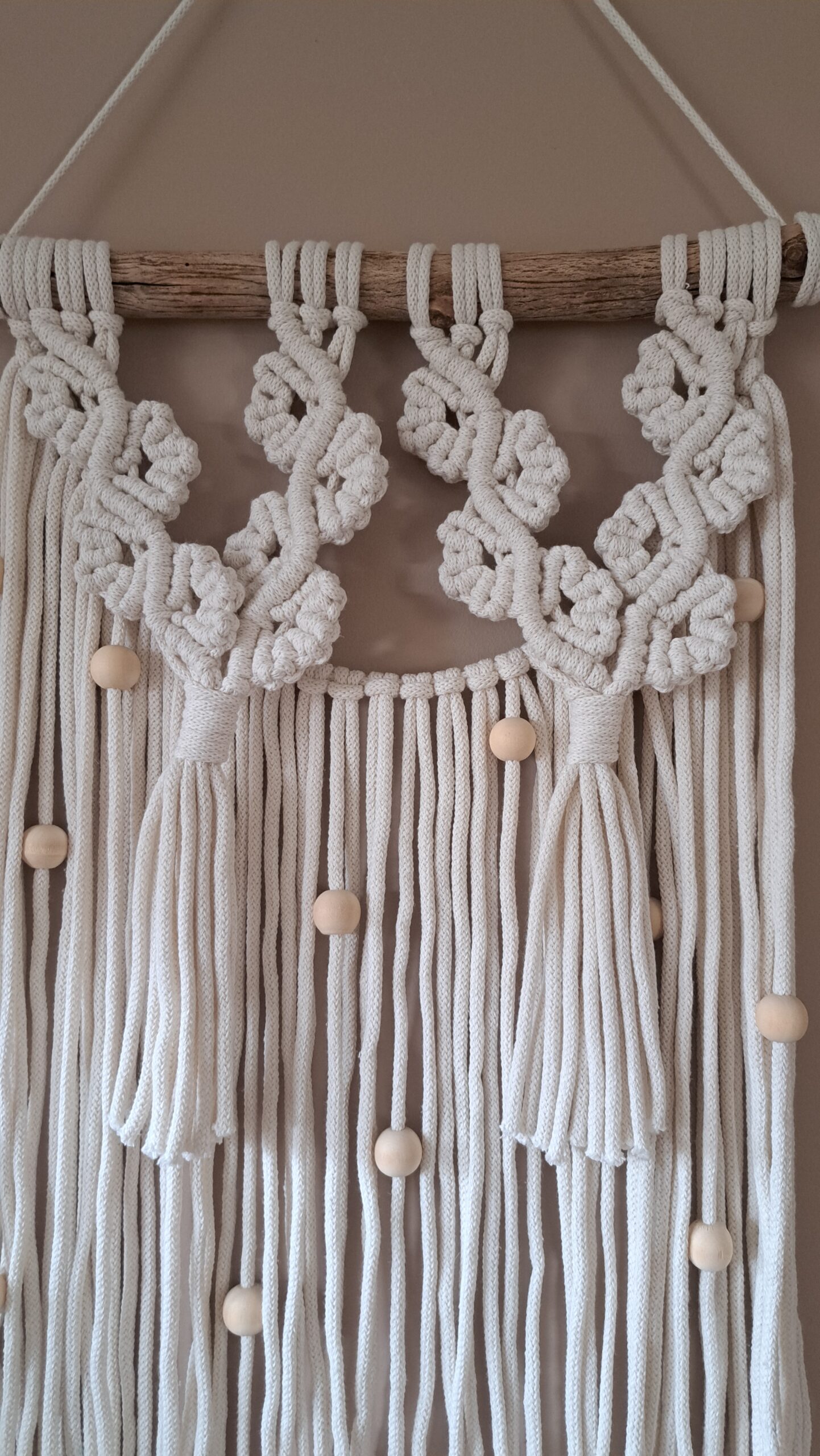 Unikalna makrama ścienna wykonana z ekologicznego sznurka bawełnianego w kolorze ecru o szerokości ok 39 cm i długości ok 55 cm. Charakterystyczny wzór z ozdobnymi splotami i licznymi drewnianymi koralikami. Zawieszona na naturalnej gałęzi, idealna do wnętrz w stylu skandynawskim, boho lub rustykalnym.