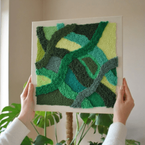 Ręce w beżowym swetrze trzymające kwadratową, teksturowaną grafikę punch needle z jaskrawymi odcieniami zieleni stylu boho. W tle fragment monstery i ciepłe, naturalne światło.