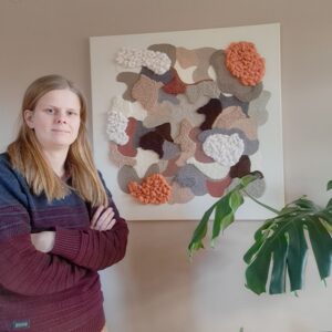 Ręcznie tkany obraz punch needle Fagus Cuprea w stylu boho. Nowoczesna dekoracja ścienna 3D do salonu w kolorach ziemi. Widoczna bogata tekstura wełny i miękkie, wypukłe sploty na białym podobraziu. Artystka stoi obok obrazu.
